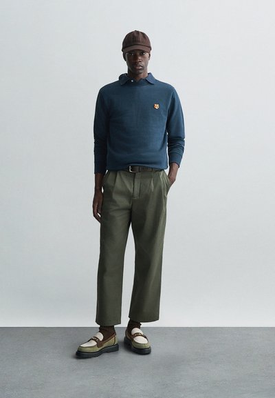 Polo cu mânecă lungă de culoare bleumarin, logo brodat, pantaloni plisati de culoare măslinie, șapcă maronie și pantofi bicolori cu accente decorative și șosete texturate.