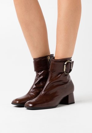 Brown patent leather ankle boots with a rounded toe, wide cuff, large round buckle, and a block heel. Zipper closure on the side.