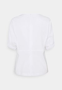 Blouse blanche à manches courtes avec poignets élastiqués et fermeture éclair au dos, vue de dos sur un fond uni.