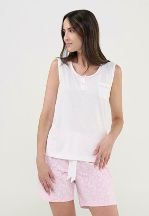 Mujer con una blusa blanca sin mangas con botones y un bolsillo, combinada con pantalones cortos rosas con estampado floral, atados con un cordón blanco.