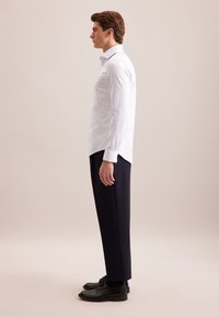 Man in een wit gestreept overhemd, zwarte broek en zwarte nette schoenen staat in profiel tegen een effen achtergrond.