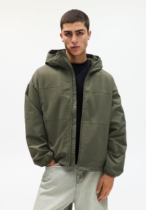 Olive green padded jacket with a hood, zip closure, and elastic cuffs. Worn over a black shirt, paired with light gray jeans.