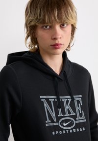 Mustapaita, jossa on suuri valkoinen Nike-logo ja teksti. Valmistettu pehmeästä kankaasta, jossa on nauhakuja ja kengurutasku.