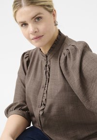 Smeđa majica s puf rukavima od teksturirane tkanine, s naboranim prednjim dijelom i detaljima s dugmadi. Okrugli izrez i skupljena tkanina na ramenima.