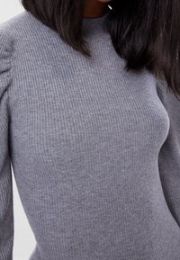 Kvinna klädd i en åtsittande grå ribbad tröja med puffärm och rak svart hår.