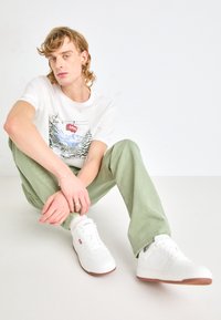 Weißes T-Shirt mit Grafikdesign in Naturmotiven, grüne Hose und weiße Sneakers mit roten Akzenten. Die Sneakers haben eine strukturierte Sohle.
