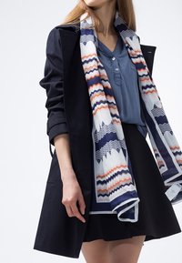 Manteau bleu marine sur une chemise bleu clair, accompagné d'une écharpe à motifs zigzag bleu marine, orange et blanc. Une jupe noire complète la tenue.