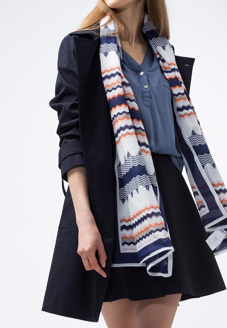 Manteau bleu marine sur une chemise bleu clair, accompagné d'une écharpe à motifs zigzag bleu marine, orange et blanc. Une jupe noire complète la tenue.