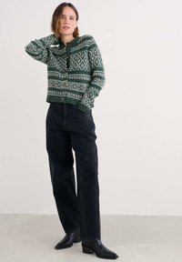 Egy nő áll, kezét a nyakára téve, zöld mintás gombos kardigánt visel, fekete bő szárú farmert és fekete bokacsizmát.