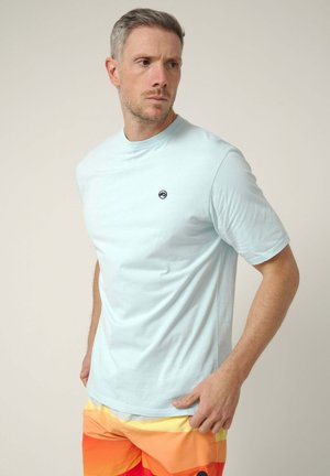 Hombre con cabello corto lleva una camiseta azul clara con un pequeño logo, pantalones cortos a rayas naranjas y amarillas, de pie contra un fondo liso, mirando hacia la izquierda.