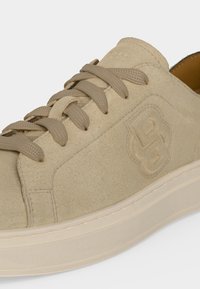 Baskets en daim beige avec lacets assortis, logo embossé sur le côté et semelle épaisse crème, présentées en gros plan sous un angle.