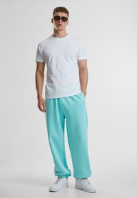 Hvit t-skjorte med korte ermer kombinert med mintgrønne treningbukser. Modell som har på seg svarte solbriller og hvite sneakers. Enkelt, casual design.