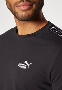 Crna pamučna majica s rebrastim okruglim izrezom koja sadrži bijeli izvezeni logo "PUMA" i kontrastnu bočnu traku s logotipima.