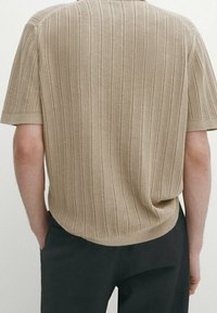 Pull en maille beige à manches courtes avec côtes verticales porté par une personne debout les mains dans les poches, associé à un pantalon foncé.