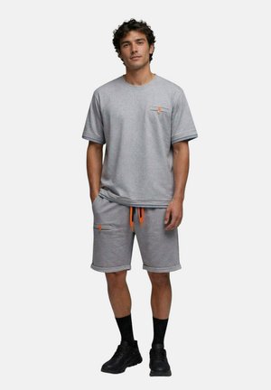 Homme debout portant un t-shirt gris à manches courtes et un short assorti avec des cordons orange, des chaussettes noires et des chaussures de sport noires, sur fond blanc.