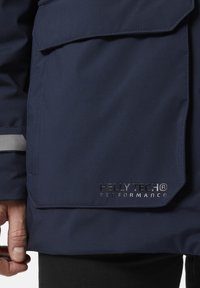 Námořnická modrá voděodolná bunda s texturovaným povrchem, vybavená velkou přední kapsou a reflexními detaily; viditelné logo "HELLY TECH® PERFORMANCE".