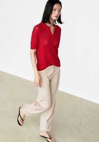 Young woman walking indoors wearing a red short-sleeve knit top, beige pants, and black flip-flops on a light floor.