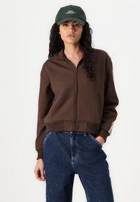 Brun zip-up sweatshirt med hög krage, ribbade muddar och en accent på vänster ärm. I kombination med löst sittande mörkblå jeans.