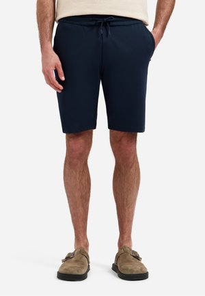 Man met marineblauwe shorts met trekkoord en beige suède instapschoenen, staand tegen een effen lichte achtergrond.