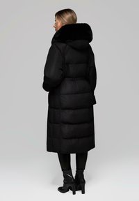 Giacca a vento nera con colletto in pelliccia nera, design trapuntato, lunghezza lunga e vita elastica. Indossata con pantaloni neri attillati e stivali con tacco.