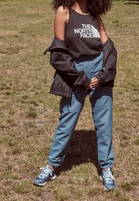 Camiseta sin mangas negra con logo blanco, combinada con pantalones jogger azul cielo y una chaqueta negra. El calzado son zapatillas azules con acentos claros.