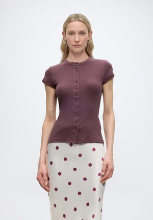 Femme blonde portant un haut prune ajusté à manches courtes avec des boutons et une jupe blanche à gros pois prune, debout sur un fond clair.