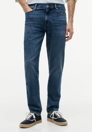 Mann trägt blaue Jeans und marineblaue Leinenschuhe mit weißen Schnürsenkeln, steht mit einer Hand in der Tasche.