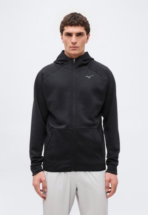 Muž v černé mikině na zip s kapucí, stojící před jednobarevným bílým pozadím, ruce uvolněné podél těla.