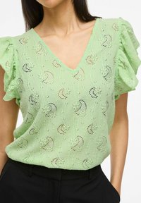 Blusa verde chiaro a maniche corte con scollatura a V, tessuto texturizzato con motivi floreali ricamati e maniche arricciate. Abbinata a pantaloni neri.