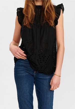 Femme portant un chemisier noir sans manches avec des détails œillets et un jean bleu, debout, une main sur la hanche et l'autre détendue.