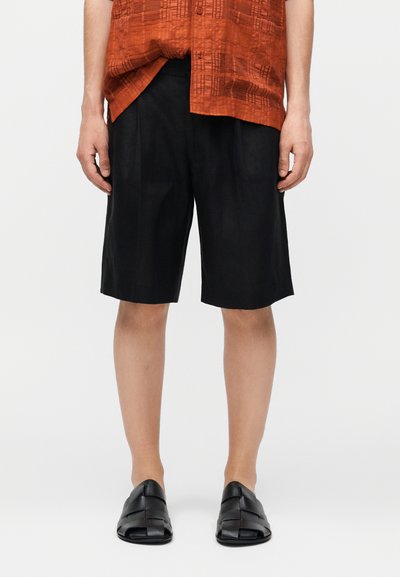 Homme portant un short noir mi-genou, des chaussures noires tissées sans lacets, et une chemise orange à manches courtes à motifs, debout droit.