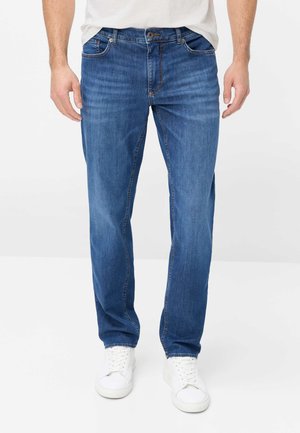 Mann trägt enge blaue Jeans und weiße Sneakers, steht vor einem einfachen weißen Hintergrund.