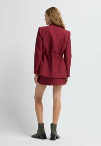 Blazer rosso con texture e vita regolabile, abbinato a una mini gonna coordinata e stivali alla caviglia verdi con tacco quadrato.