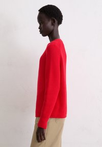 Rode gebreide trui met lange mouwen en geribbelde manchetten, met een ronde halslijn en een gestructureerd oppervlak, gecombineerd met beige broeken.