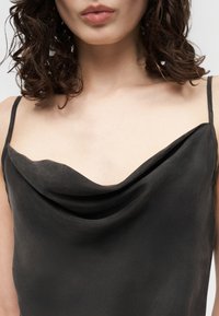 Kvinne med krøllete brunt hår iført en svart satengkjole med halssmykke og tynne skulderstropper mot en nøytral bakgrunn.
