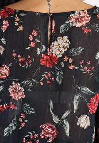 Blouse florale avec un fond sombre, ornée de fleurs rouges, blanches et vertes. Fermeture au dos avec un petit détail en trou de serrure et une texture subtile.