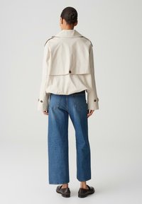 Giacca beige leggera con uno spessore sul retro e bottoni, abbinata a jeans blu a gamba larga e scarpe slip-on scure.