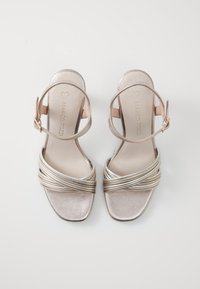 Las sandalias de tacón en plata metálica y dorada presentan un diseño con tiras. Textura suave con punta cuadrada y hebilla en el tobillo ajustable.