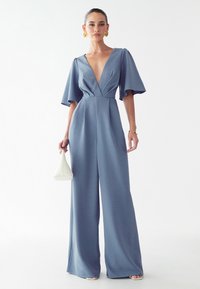 Modra kombinezona z globokim V-izrezom, kratkimi flared rokavi in širokimi hlačnicami. Gladka tekstura, v kombinaciji z belo geometrijsko torbico.