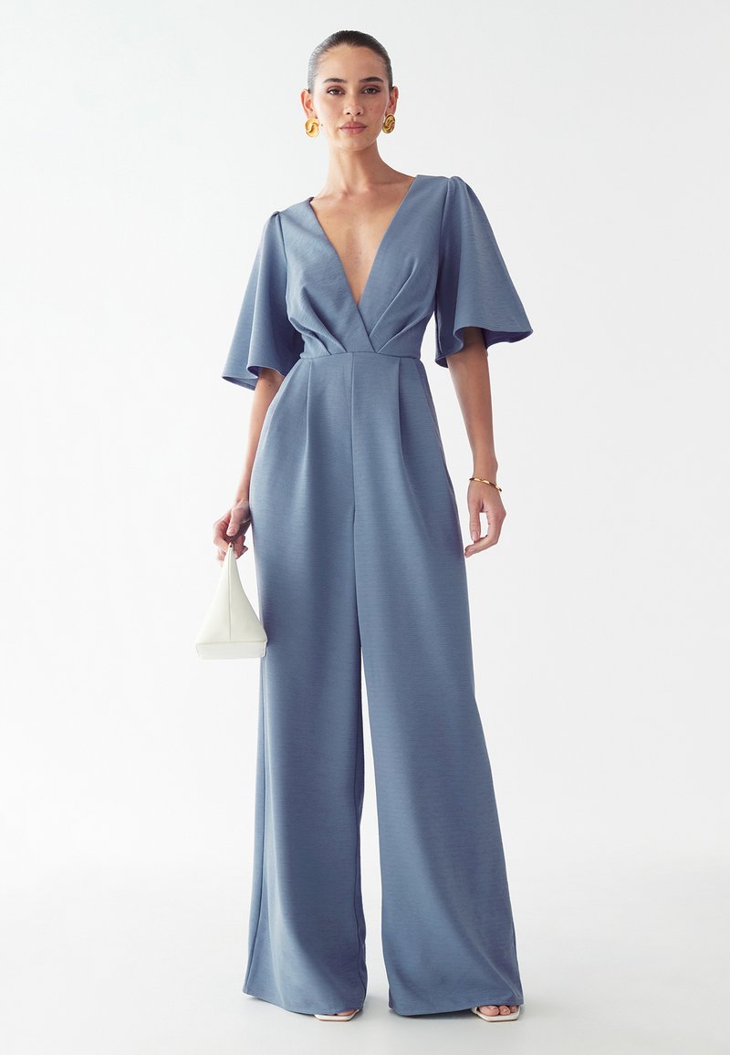 Modra kombinezona z globokim V-izrezom, kratkimi flared rokavi in širokimi hlačnicami. Gladka tekstura, v kombinaciji z belo geometrijsko torbico.