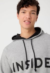 Sudadera con capucha gris, con cordones negros y una capa inferior negra en la capucha, que presenta un atrevido texto negro "INSIDE" en el pecho.