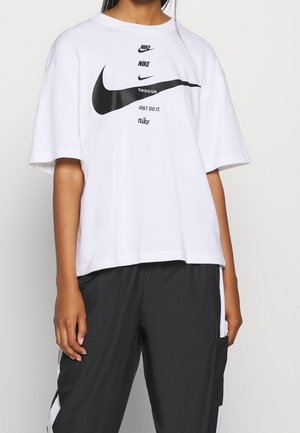 Person wearing a white Nike T-shirt with multiple logos and "Just Do It" text, paired with black and white sporty pants.