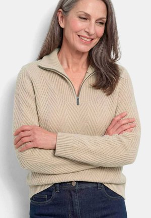 Femme aux longs cheveux gris portant un pull zippé beige texturé et un jean bleu foncé, souriante, les bras croisés.