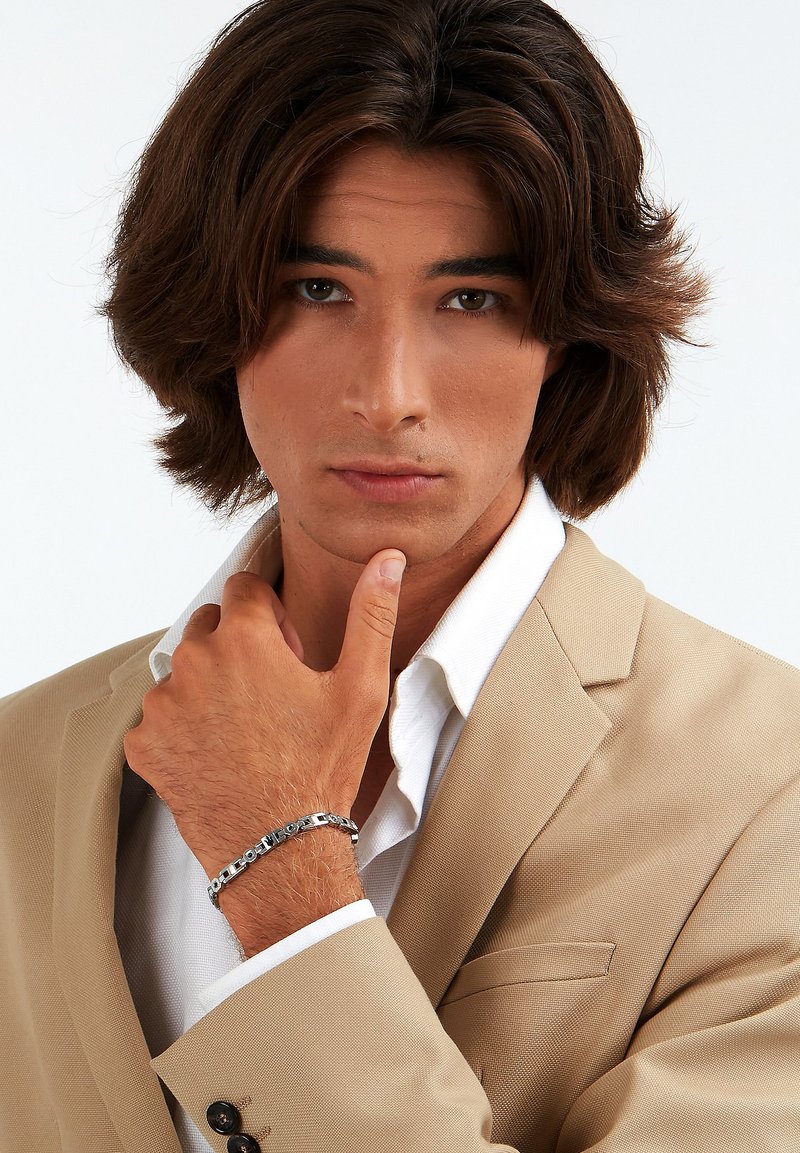 Beige colbert over een witte blouse, met een gestructureerd patroon. Zilveren armband met tekstaccenten siert de pols. Lange, donkere haren.