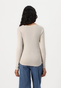 Cămașă bej cu mâneci lungi, cu decolteu rotund și tiv curbat, asortată cu pantaloni denim lejeri de culoare albastră. Textura este moale și ajustată.