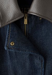 Veste en denim foncé avec col en cuir brun texturé et détails apparents en laiton sur la fermeture éclair et les boutons-pression.