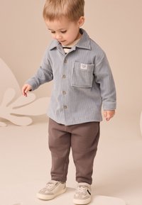 Chemise en velours côtelé bleu clair avec boutons marron et poche poitrine, assortie à un pantalon marron foncé et des baskets claires, sur un enfant debout.