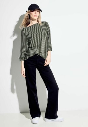 Haut ample rayé vert et blanc, pantalon noir évasé et baskets blanches ; modèle portant une casquette noire, debout contre un fond clair.