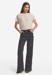 Kvinna i en lös beige blus instoppad i högmidjade, vida svarta jeans med svart bälte, stående med händerna i fickorna.