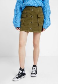 Gonna mini in denim verde oliva con bottoni frontali e grandi tasche, abbinata a un maglione chunky azzurro e sneakers alte nere.